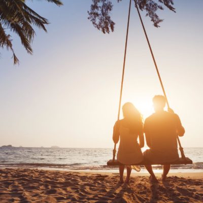 A lady sits next to a callboy on a swing right on the beach and watches the sunset
