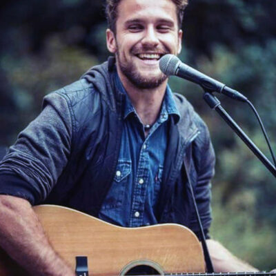 ein junger Mann mit braunen Haaren lächelt mit einer Gitarre und singt