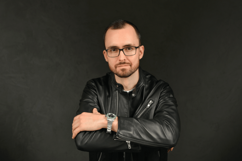 Callboy Alexander stands confidently against a shadowed backdrop. He has short, neatly styled hair, a well-groomed beard, and wears stylish glasses that complement his features. Dressed in a sleek black leather jacket paired with a modern wristwatch, Alexander exudes understated confidence. With his arms crossed over his chest and maintaining a neutral expression, he projects an air of calm self-assurance and quiet strength amidst the moody darkness behind him.