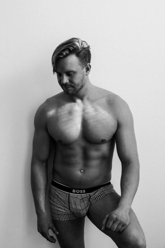 This black and white image creates a striking and mysterious portrait of Callboy Michael. The focus on his styled hair and the way light plays across his chest emphasizes both his physique and enigmatic allure, while the patterned boxer briefs add a touch of personality. The plain white wall backdrop ensures that attention remains solely on him, enhancing the overall sense of intrigue in the composition.