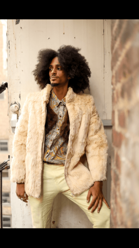 This description evokes a striking image of someone with a bold sense of style and confidence. The voluminous curly hair, similar to Callboy Leo Knights, adds to the persons distinctive look, while the cream-colored fur coat adds a touch of luxury and warmth. Paired with a patterned shirt and light-colored pants, the ensemble suggests a fashionable yet relaxed vibe. The brick wall backdrop provides an urban feel that contrasts well with their sophisticated outfit. Overall, this scene captures an intriguing blend of textures and patterns, enhanced by the individuals poised stance and thoughtful gaze into the distance.