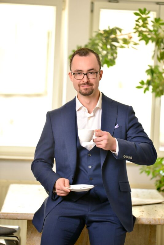It sounds like youre describing a scene for a photo, illustration, or character profile. Here’s a possible descriptive paragraph based on your prompt: Callboy Alexander exudes quiet confidence as he sits casually atop a polished counter. Dressed in an impeccably tailored blue suit and crisp white shirt, he holds a delicate white teacup and saucer with practiced ease. His short hair is neatly styled, complementing his well-groomed trimmed beard—a look further refined by the sleek glasses perched on his nose. Sunlight pours through expansive windows behind him, casting soft highlights across the room and illuminating flourishing green plants that add lively contrast to the modern decor. If you need this as part of a creative writing piece or would like an AI-generated image of this scene (if using compatible tools), just let me know!.
