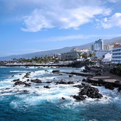 Die Küstenlandschaft von Mauritius präsentiert eine malerische Mischung aus natürlicher Schönheit und urbanem Charme. Die felsigen Ufer treffen auf die kaskadierenden Meereswellen und schaffen eine ruhige und zugleich dynamische Umgebung. Entlang der Küste erheben sich stolz Gebäude unter einem teilweise bewölkten Himmel, deren Design das tropische Flair der Region unterstreicht. In der Ferne bilden die Berge eine majestätische Kulisse, die der Landschaft Tiefe verleiht. Verstreut wiegen sich Palmen sanft im Wind und erinnern an idyllische Postkarten, die die Essenz des Paradieses einfangen. Diese lebendige Landschaft wird durch das warme Sonnenlicht, das durch die Wolken fällt und verspielte Schatten auf Land und Meer wirft, noch verstärkt.