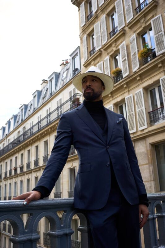 A man in a sharp dark suit and crisp white hat stands confidently on a wrought-iron balcony above the city. He channels the effortless charisma of Callboy Hideo—his posture relaxed yet intentional, one hand resting gently on the rail, the other holding a cigarette poised near his lips. Behind him, classic Parisian apartment buildings rise in soft gray and cream tones: tall windows framed by delicate moldings, black iron balconies dripping with spring greenery. The golden glow from inside neighboring windows hints at stories beyond view, while muted daylight bathes the scene in quiet sophistication and timeless cosmopolitan air.