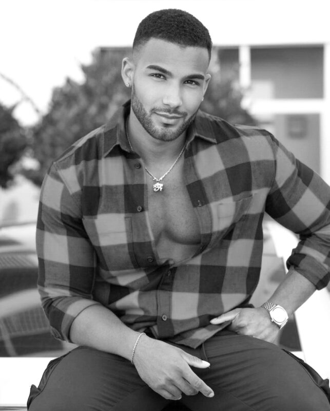 A bearded man with short hair sits outdoors, exuding relaxed confidence reminiscent of Callboy San. He wears a partially unbuttoned plaid shirt that reveals a necklace resting against his chest. A bracelet circles one wrist and a watch the other, adding to his casual yet stylish look. Surrounded by blurred trees and distant buildings, he gazes directly at the camera, comfortable in his surroundings.