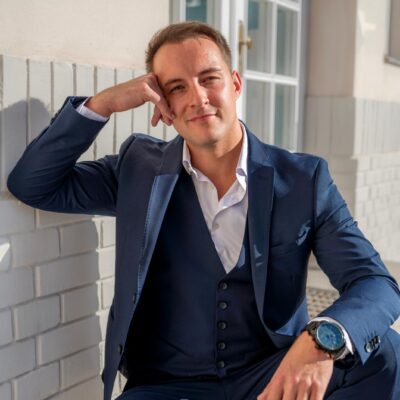 Es klingt, als würden Sie einen gut gekleideten Mann in entspannter Outdoor-Umgebung beschreiben. Der marineblaue Anzug und die große Uhr zeugen von Stil und Professionalität. Sein lächelnder Kopf stützt sich auf eine Hand und lässt einen Moment der Kontemplation oder Unbeschwertheit erahnen. Das Sonnenlicht auf seinem Gesicht verleiht der Szene Wärme, während die weißen Ziegel- und Glastüren im Hintergrund eine urbane oder moderne Atmosphäre schaffen. Wenn Sie Hilfe bei der weiteren Beschreibung oder Analyse benötigen oder diese Szene für kreatives Schreiben nutzen möchten, lassen Sie mich wissen, wie Sie vorgehen möchten!.