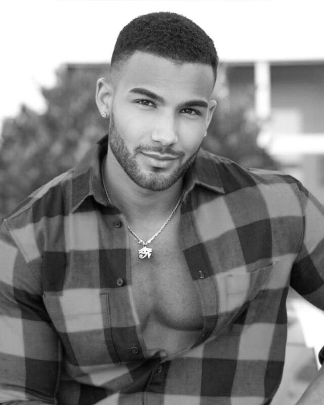 Certainly! Here’s a description of your black and white photo suitable for sharing or artistic purposes: --- A man with short hair and a neatly trimmed beard sits outdoors, exuding relaxed confidence. His plaid shirt is partially unbuttoned, revealing a simple necklace that adds to his casual charm. Looking directly at the camera with a subtle, inviting smile, he embodies the classic allure of Callboy San. The monochrome palette emphasizes the contrasts in light and shadow, highlighting his features and the textures of his attire. --- If you’d like an AI-generated image based on this description or need further refinement for another use (profile, writing project), let me know!.