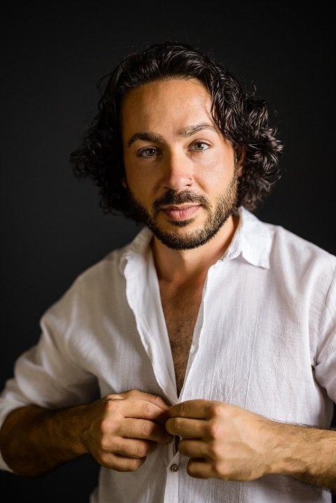 It sounds like youre describing a cinematic or stylish portrait of a man: he has curly dark hair and a beard, is standing against a dark background, and wears an open-collared white shirt. He looks towards the camera, adjusting his shirt, which may suggest confidence or casual elegance. If you’d like to use this description for writing, characterization, or visual reference purposes, here’s an enhanced version: --- A man with curly dark hair and a well-groomed beard stands confidently before a deep shadowy backdrop. Dressed in an open-collared white shirt that contrasts sharply with the darkness behind him, he meets the cameras gaze directly. One hand lifts to adjust his collar thoughtfully, lending him an air of relaxed self-assurance. --- If you want something specific (for example—a prompt for generating images or more poetic language), let me know your intended use!.