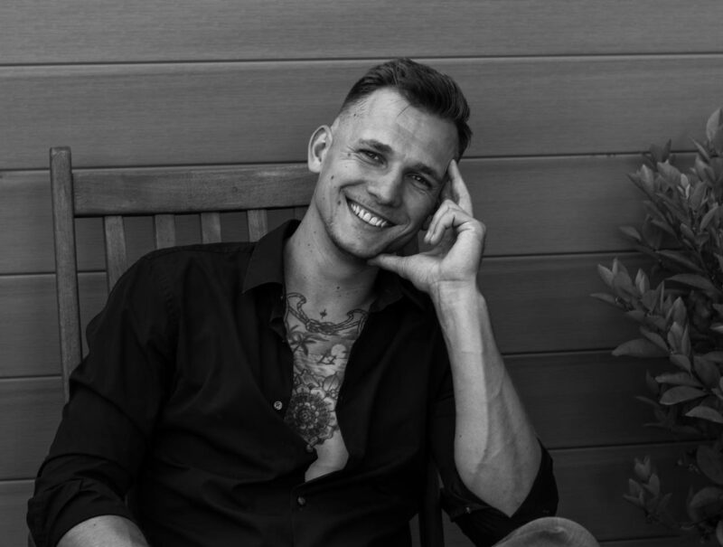 It sounds like you are either describing a portrait photograph or seeking to generate an image based on this description. Here’s how I interpret your description: A man with short hair and a warm, friendly smile sits comfortably on a wooden chair. He has a noticeable chest tattoo that is partially visible because of his open dark button-down shirt. He leans slightly forward, resting his head thoughtfully on one hand in a relaxed pose. The setting features an elegant paneled wall behind him, adding depth to the composition; beside him are some leafy green plants that introduce an element of freshness and color contrast. If you need suggestions for posing, photography tips, or want this rewritten as part of an art prompt or story scene, let me know!.