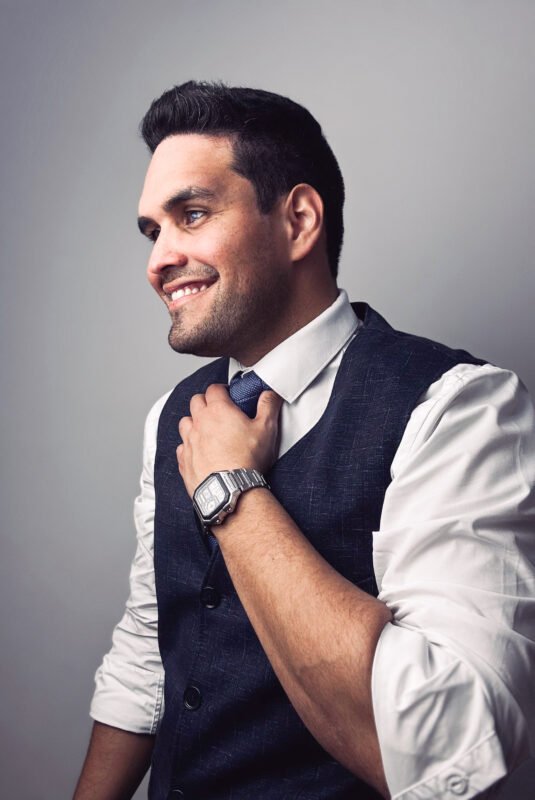 The scene you describe suggests someone who is confident and perhaps preparing for a formal occasion or an important event. The man’s attire—vest, dress shirt, watch, and tie—implies professionalism or a polished sense of style. His smile and the act of adjusting his tie while looking to the side create an impression of approachability and readiness, possibly captured in a candid moment before heading into a meeting, interview, or social gathering. The plain light background helps put full focus on him without distractions. If you need this description refined for creative writing, professional use (like LinkedIn), or as part of photo metadata/captioning, let me know how youd like it tailored!.