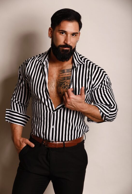 A confident man stands before you. He has a dark, well-groomed beard and styled hair, exuding a bold charm. He wears a black-and-white striped shirt thats partially unbuttoned, showing off an intricate chest tattoo beneath. One hand is casually tucked into his pocket, while the other grips the edge of his open shirt, drawing attention to his tattoo and adding to his self-assured pose.