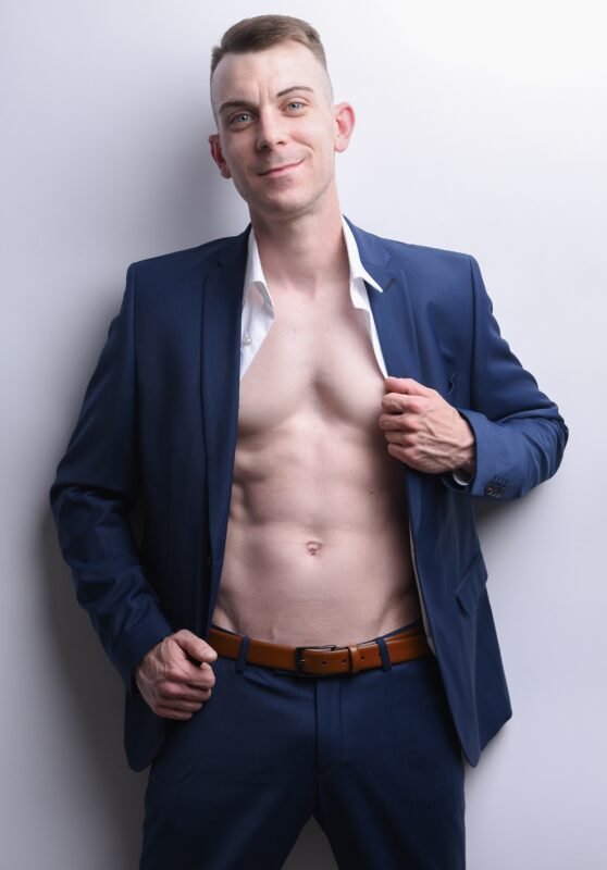 Dennis steht selbstbewusst vor einem schlichten, hellen Hintergrund. Er trägt einen eleganten blauen Anzug; die Jacke ist offen, und sein frisches weißes Hemd hängt aufgeknöpft und gibt den Blick auf seine nackte Brust und seine definierten Bauchmuskeln frei. Seine entspannte Pose und sein selbstbewusster Ausdruck strahlen Charisma aus, während der Kontrast zwischen dem maßgeschneiderten Anzug und dem freiliegenden Oberkörper sowohl auf Eleganz als auch auf Anziehungskraft hindeutet.