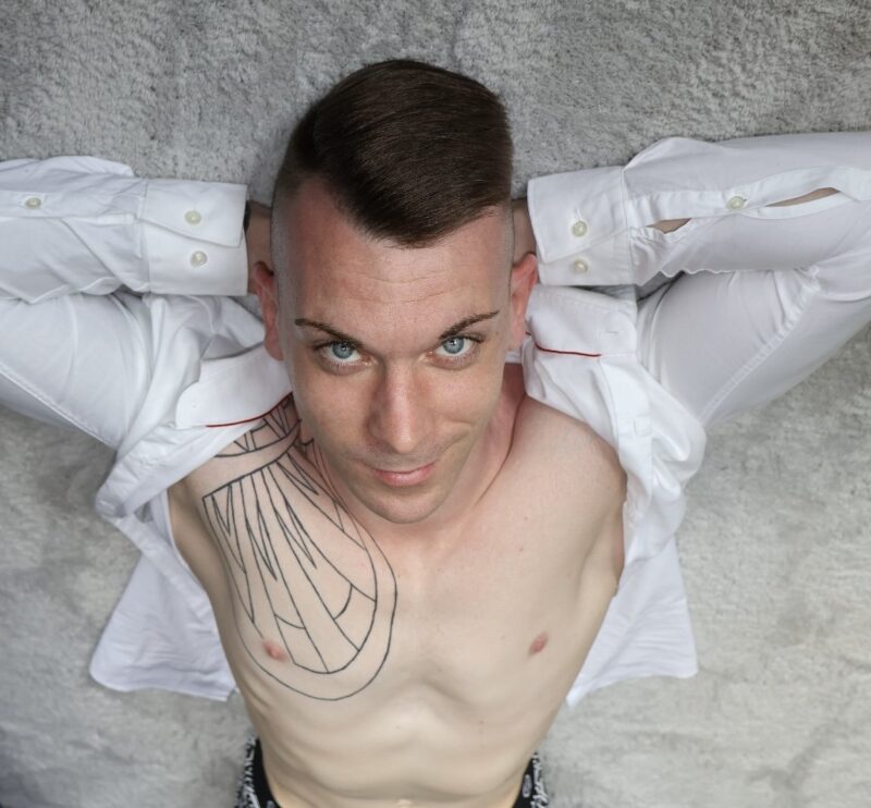 You’ve provided a description of an image: a man, “callboy Dennis,” lies on his back on a light gray carpet, looking up at the camera. He’s wearing an open white shirt and has a large geometric tattoo on his chest and shoulder, with his arms relaxed behind his head. If you have any questions or would like help expanding this description (for example, for creative or technical purposes), please let me know how I can assist you!.