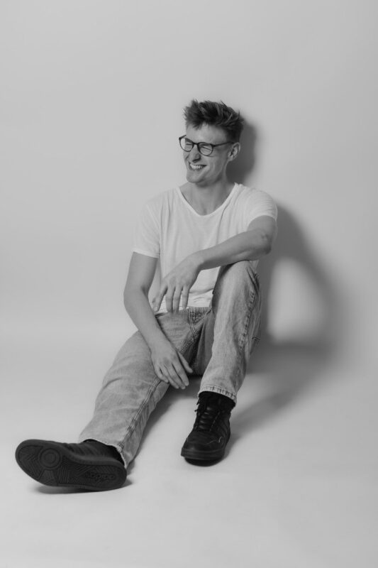 Certainly! Here’s a descriptive text for your scenario: A young man—Callboy Moritz—sits casually on the floor, his back resting against a smooth, unadorned wall. He wears simple black-framed glasses and a plain white t-shirt that contrasts with his classic blue jeans and black shoes. His posture is relaxed: one knee up, an arm draped loosely over it, as he smiles warmly, eyes turned thoughtfully to the side. The photograph is rendered entirely in black and white, emphasizing light and shadow across his features and clothing against the minimalist backdrop. If you need this description tailored further or written in another style (e.g., for visual artists or photographers), let me know!.
