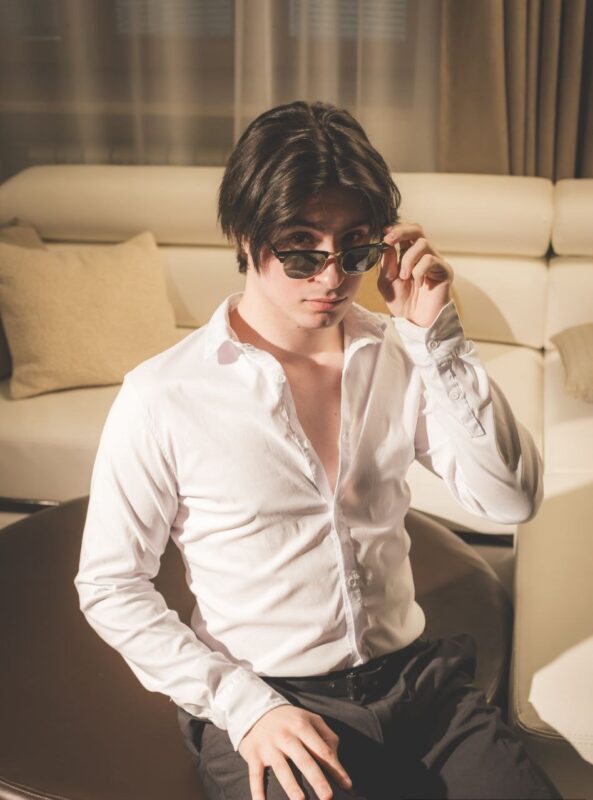 Luka, a bi callboy with medium-length dark hair, sits casually on a round ottoman in the middle of a sunlit living room. He sports a crisp white button-down shirt—slightly unbuttoned at the top—and fitted black pants. With an effortless coolness, Luka tilts his sunglasses down the bridge of his nose, gazing over them with one eyebrow raised in playful intrigue. Soft sunlight pours through broad windows behind him, warming the modern furnishings and casting gentle highlights through his hair as he strikes this inviting pose.