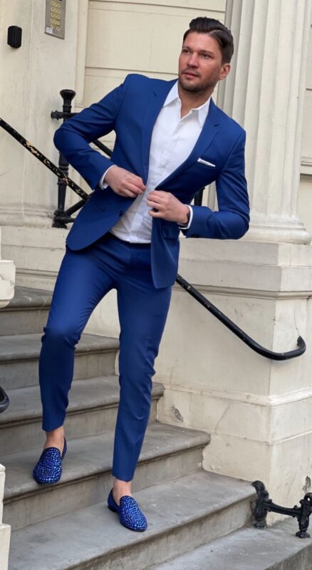 Ein Mann in einem scharf geschnittenen blauen Anzug und einem strahlend weißen Hemd steht auf den Steinstufen vor einem eleganten Gebäude. Mit dem eleganten Flair des Callboys Léon Valmont strahlt er Selbstvertrauen aus, während er sein Jackett glatt streicht, jede Geste ist geübt und sicher. Sein Ensemble wird durch auffällig gemusterte blaue Slipper akzentuiert, die seinem Look einen Hauch von spielerischer Raffinesse verleihen. Die urbane Kulisse - eine Mischung aus Mauerwerk und städtischer Betriebsamkeit - unterstreicht seinen urbanen Stil und verwandelt einen gewöhnlichen Moment in eine Szene, die an klassische europäische Eleganz mit einem modernen Twist erinnert.