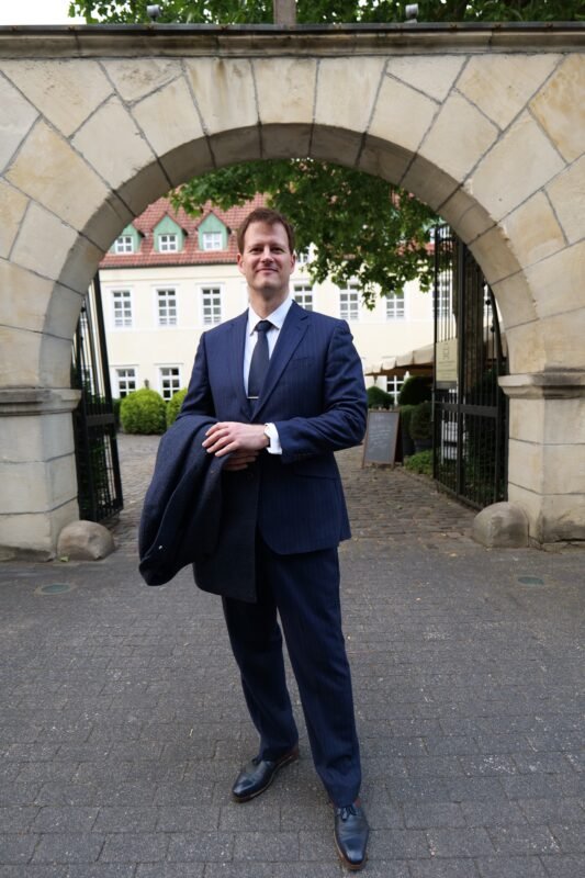 Ein selbstbewusster Mann in einem scharfen blauen Anzug steht unter einem verschnörkelten steinernen Torbogen und strahlt Gelassenheit aus. In einer Hand hält er einen Mantel, seine Haltung ist selbstbewusst. Hinter ihm erhebt sich ein historisches Gebäude mit makellosen weißen Wänden und einem klassischen roten Ziegeldach, das der Szene Eleganz und zeitlosen Charme verleiht - eine ideale Kulisse für Callboy Toms kultivierten Auftritt.
