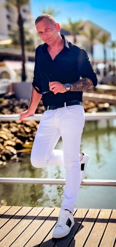 Ein Mann in schwarzem Hemd und weißer Hose steht lässig auf einem Holzdeck mit Blick auf das Wasser und nimmt eine entspannte Pose ein, die an das selbstbewusste Auftreten eines Callboys erinnert. In einer Hand hält er eine Sonnenbrille, die einen entspannten Charme ausstrahlt. Im Hintergrund wiegen sich hohe Palmen sanft neben modernen Gebäuden unter einem strahlend blauen Himmel und schaffen eine lebendige und tropische Atmosphäre am Wasser.